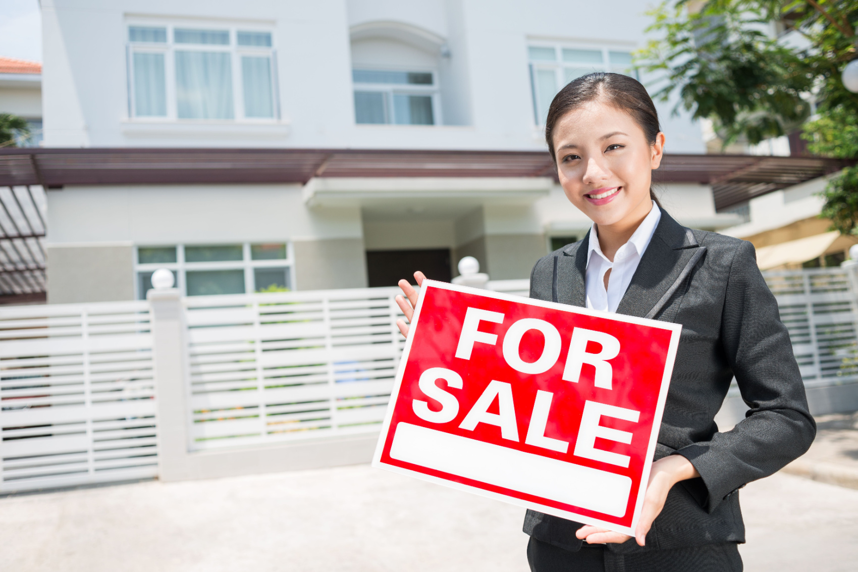 Female real estate worker holding a "For Sale" sign in front of a vacant real estate property.